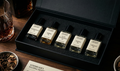 Set of five small bottles of perfume in a black box on a wooden surface with a glass of whiskey and a bowl of sweet spices.